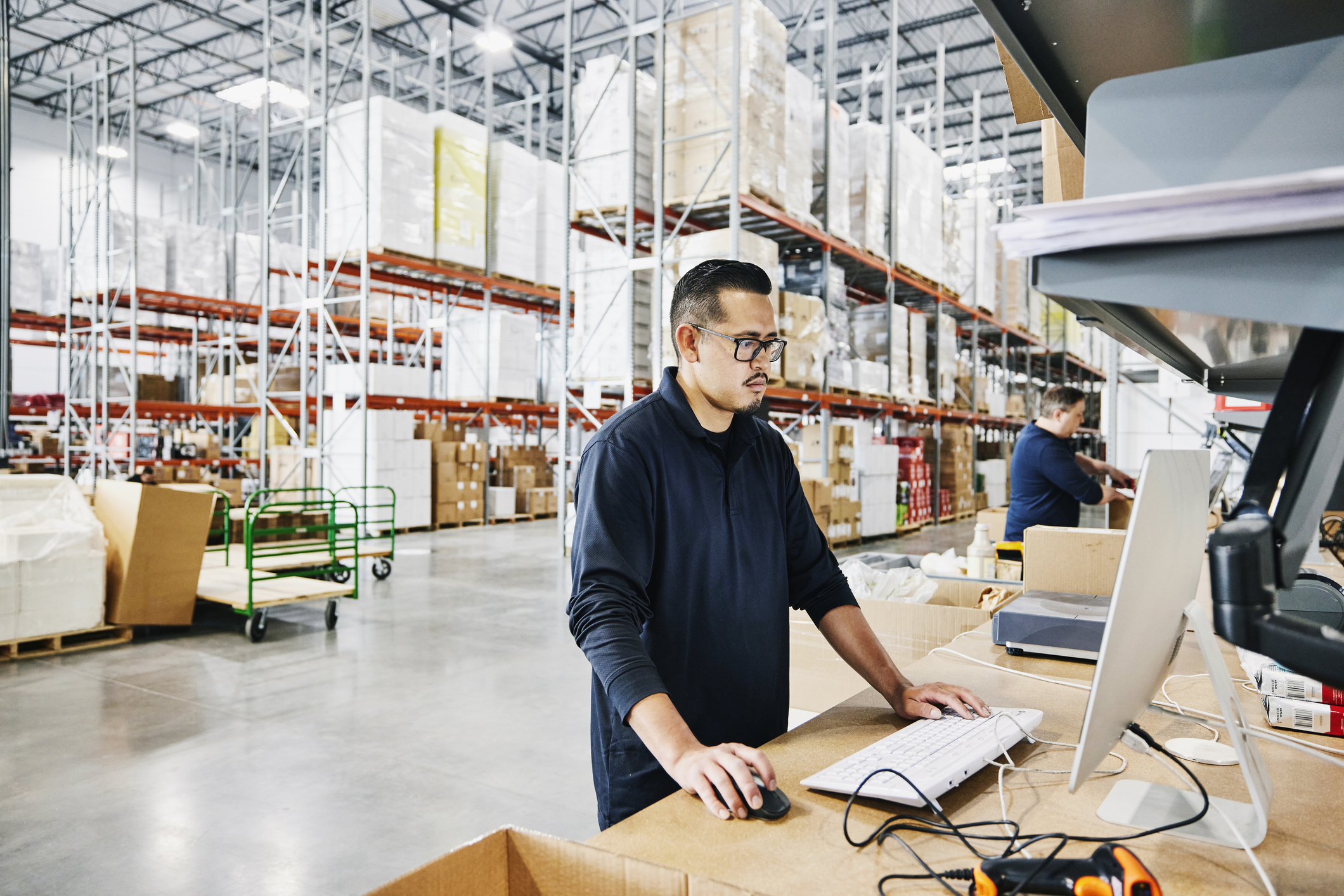 Two men working on computers inside a warehouse stocked with inventory and boxes.