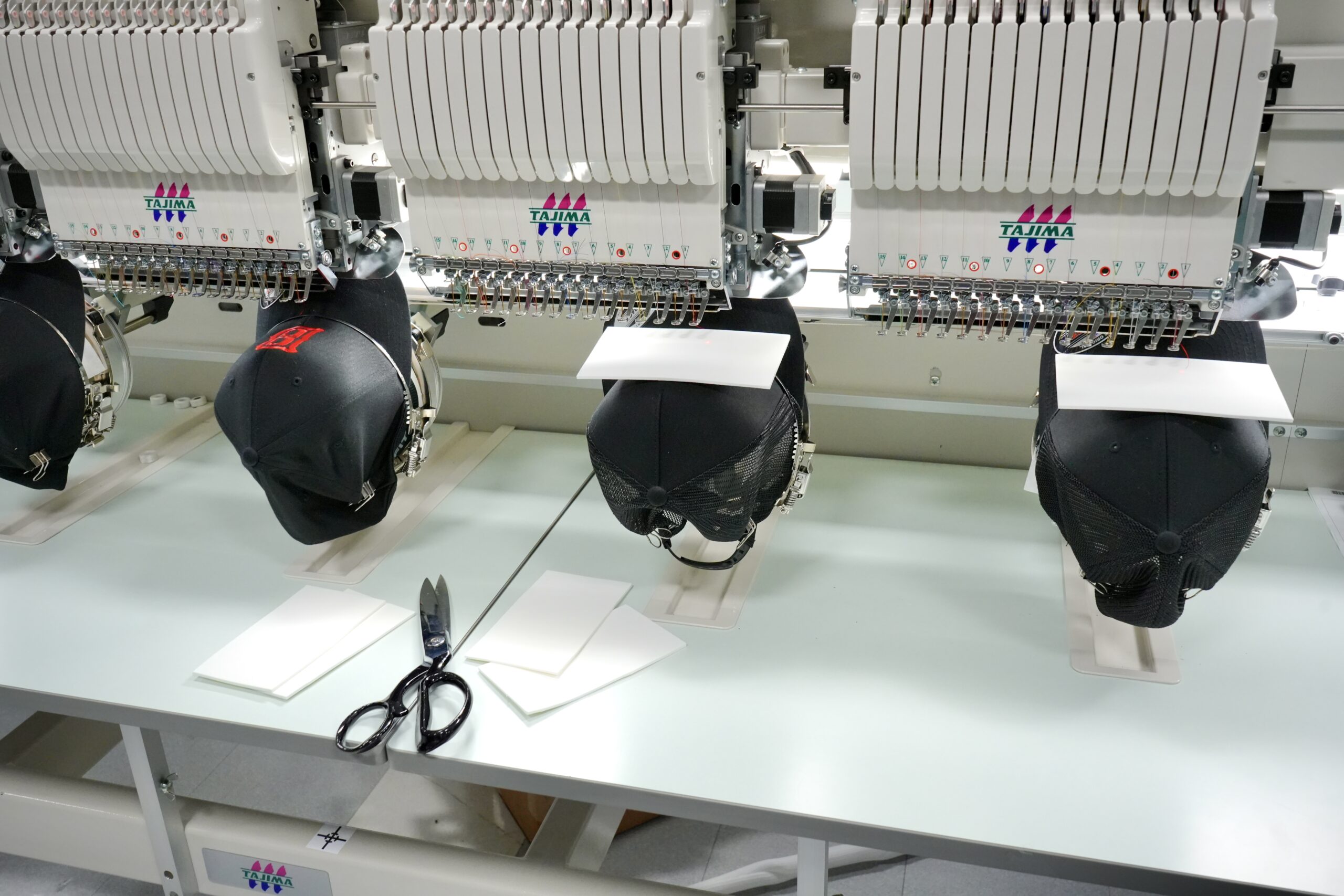 Four black caps on an embroidery machine, two with red thread and two with white foam overlay
