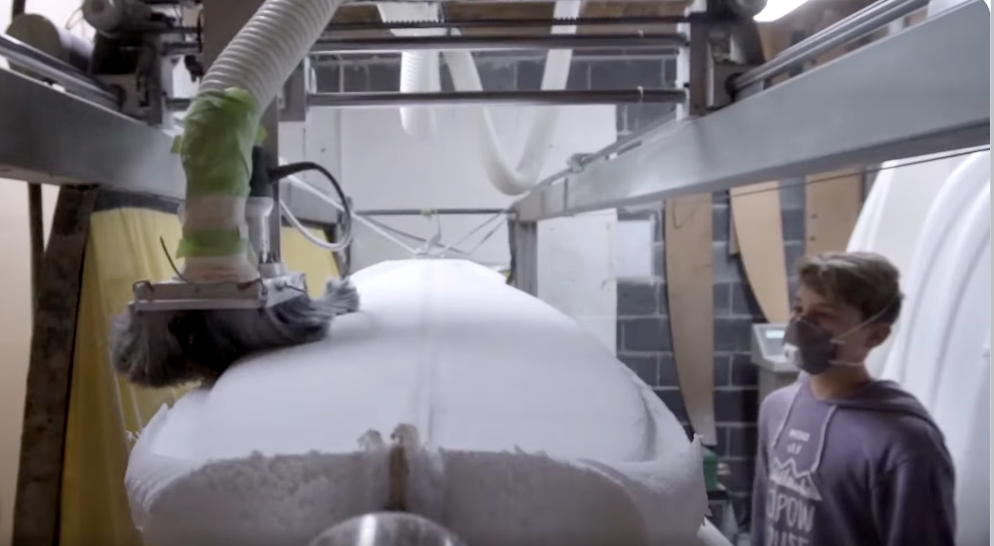 A person standing next to a blank white surfboard being customized