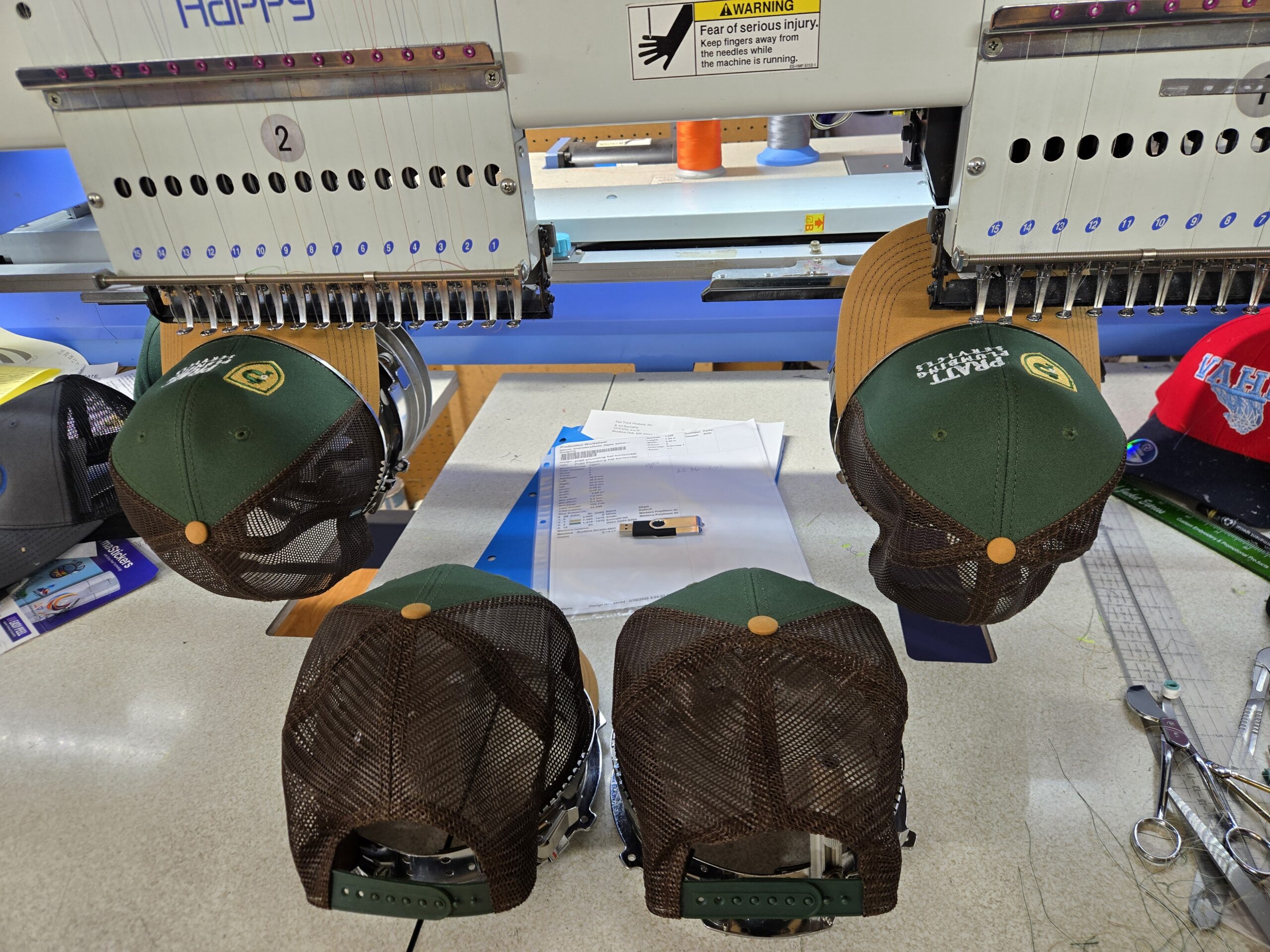 Two black hats on an embroideyr machine, with two additional black hats between them on a desk