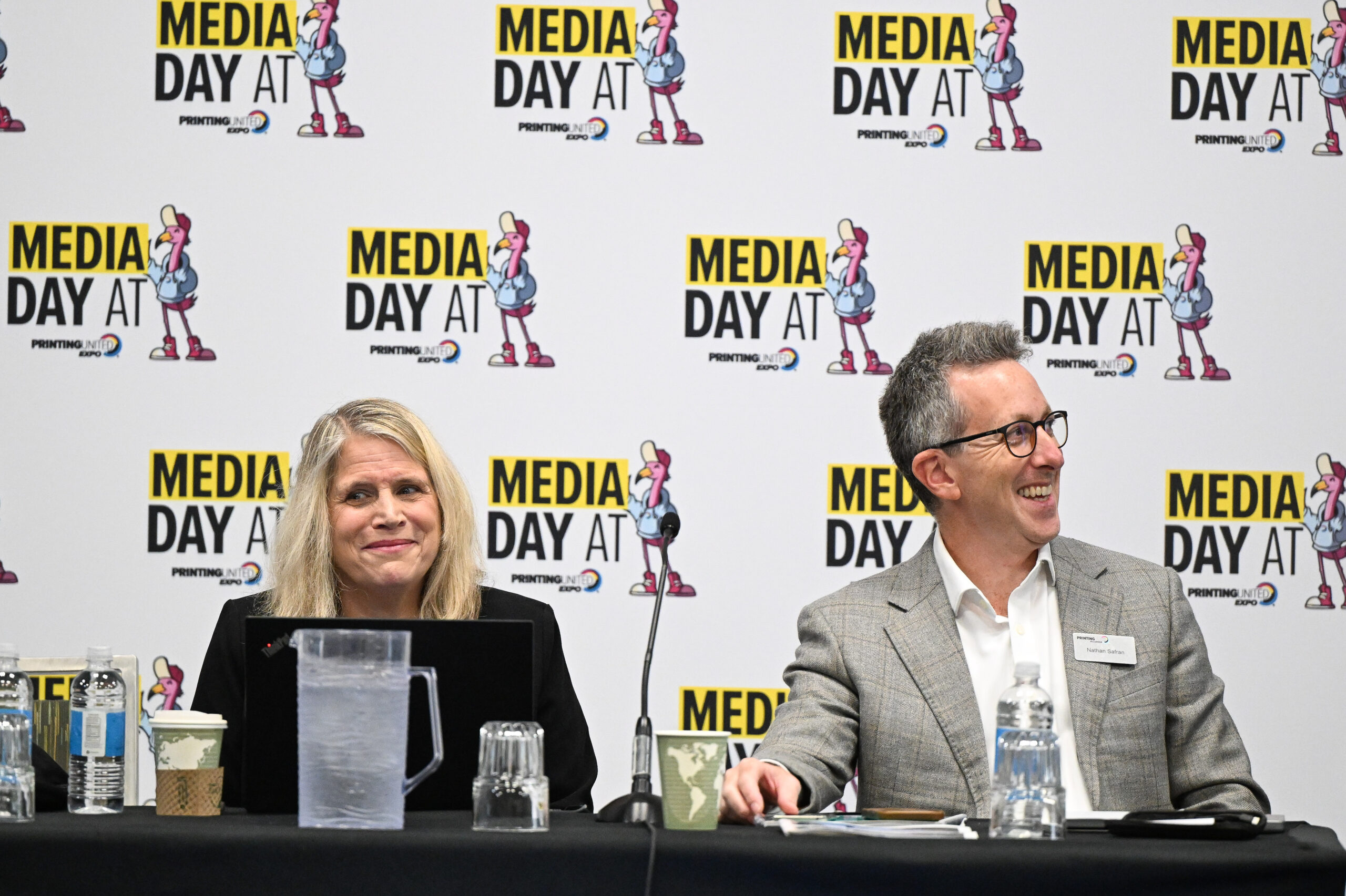 A woman and a man both wearing suits sit at a press table with microphones and a step and repeat background