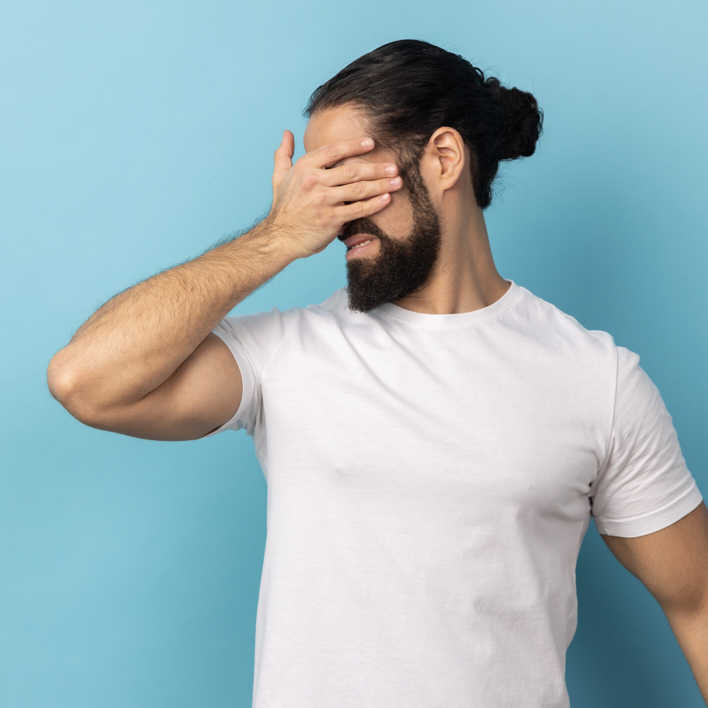Don't want to look at this. Portrait of scared man with beard wearing white T-shirt standing, covering eyes with hand, refusing to watch. Indoor studio shot isolated on blue background.