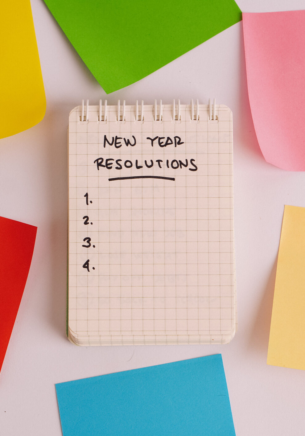 A notepad featuring the handwritten phrase &ldquo;New Year&rsquo;s Resolution&rdquo; at the top of the page, symbolizing a fresh start. The notepad is open surround with multicolored adhesive notes, inviting viewers to start planning their goals. The simple, personal feel of handwriting emphasizes reflection and commitment, ideal for content related to New Year&rsquo;s goals, personal growth, and wellness.